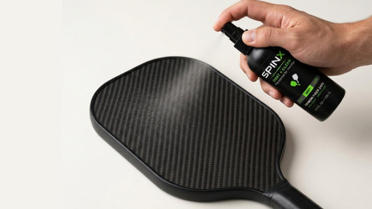 A hand spraying SPINX COAT + CLEAN onto the face of a black carbon-fiber pickleball paddle